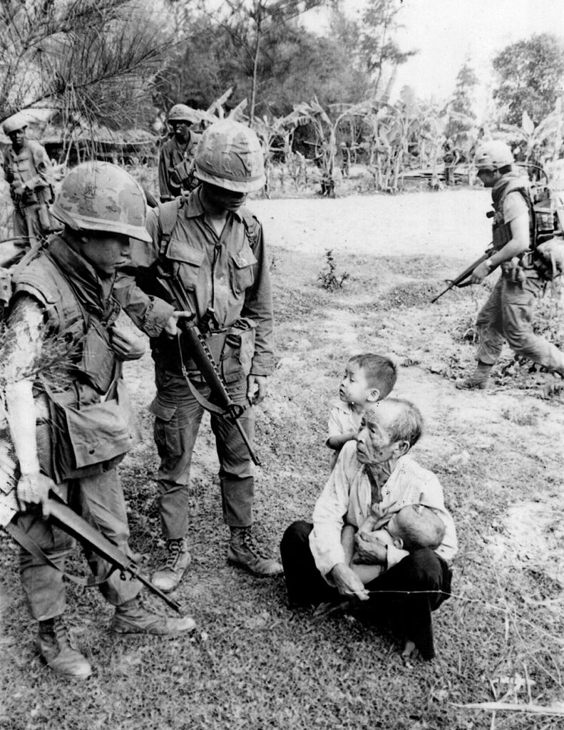 American soldiers interrogate an old man and two children, one an infant at the time of the My Lai massacre. Two African-American soldiers are present in the background. Photograph by Ronald L. Haeberle, 1968, source: Wikimedia Commons